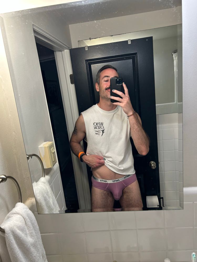 mirror selfie of Kyle wearing a white cut-off Betty Who tee and pink briefs