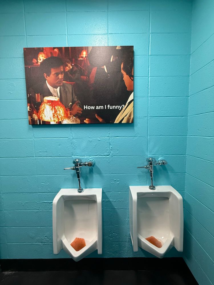 picture of two urinals with a large My Cousin Vinny poster hanging above them that reads “How am I funny?”