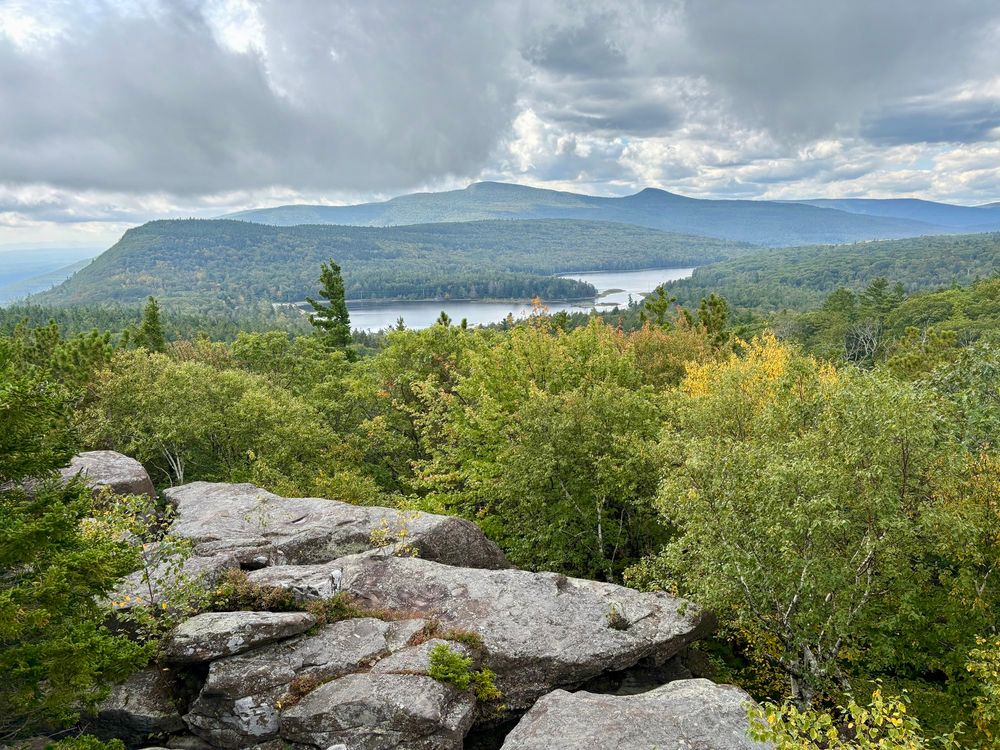 Photograph looking from a rocky ledge across forest to a lake in the middle distance. Forested mountains rise beyond.