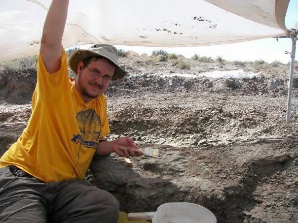 Photograph of my much younger self, in yellow t-shirt and gray Tilley hat, posing beneath a sun-tarp midway through scraping at a shelf of mudstone in the Utah desert.