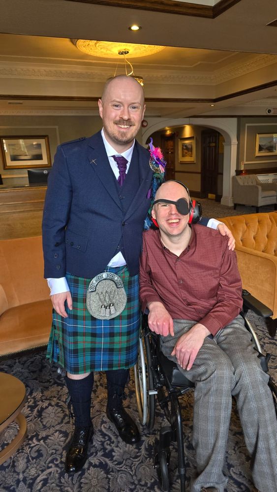 Image depicts two inordinately handsome men in the lobby of the Kingsmills Hotel during a wedding. On the left, standing, the groom: a man in a blue suit jacket and a green-and-blue tartan kilt. On the right, myself, sat in my wheelchair with my eyepatch on looking like Billy Corgan if he took up piracy during the Mellon Collie years.