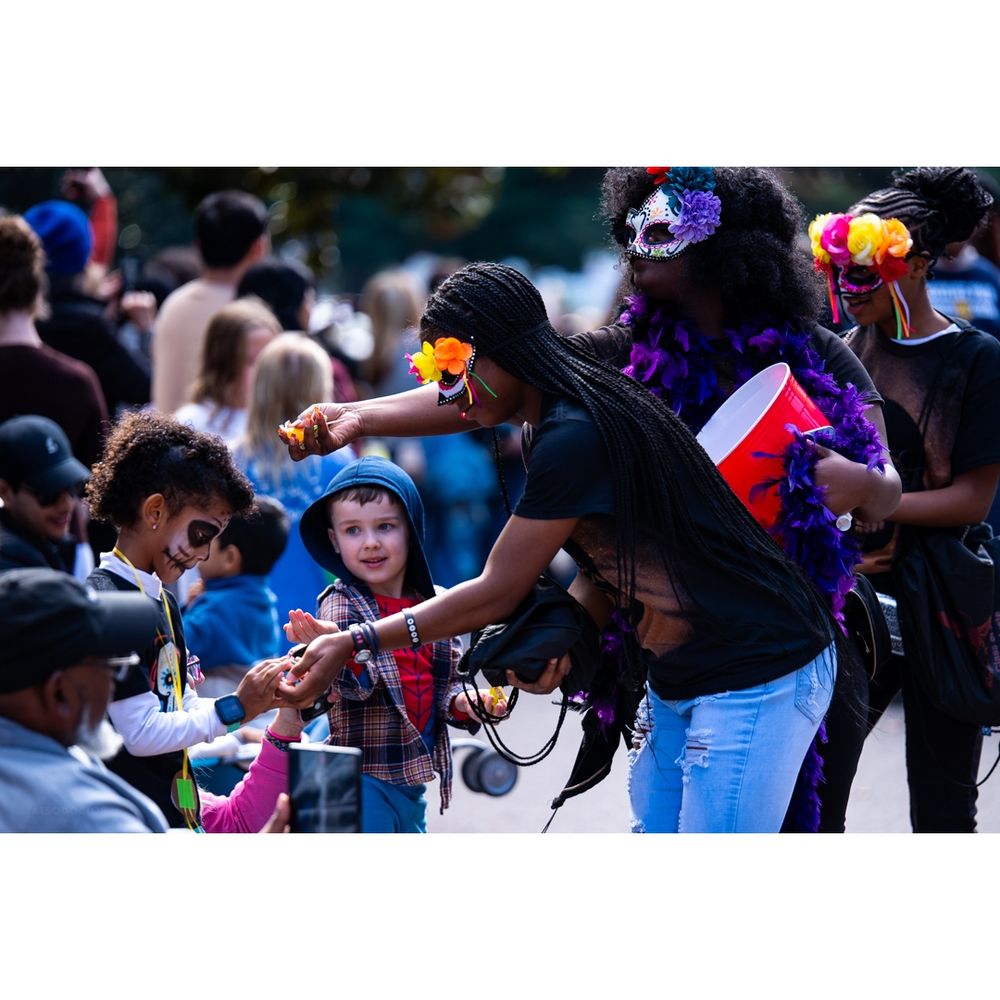 Color. Day. Several children on the left. One, in profile, wearing skull makeup. The children have their hands out. On the right, three teenagers in dark shirts, skull makeup and half-masks hand candy to the children's outstretched hands.