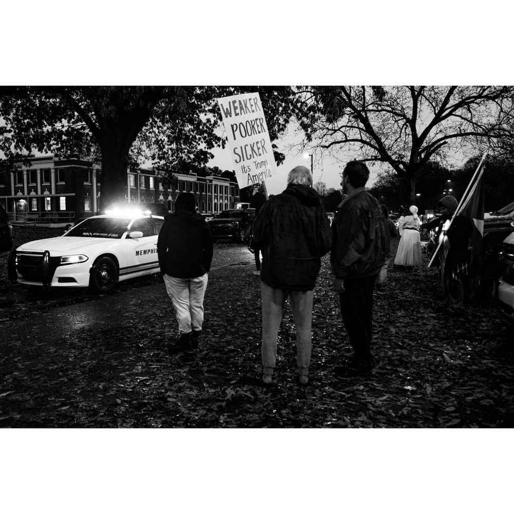 B&W. Night. Rain and fallen leaves. In the background, the black skeletons of large trees and, to the left, a county administration building. Mid-ground, left: a police car, emergency lights on. Foreground, center: The backs of a group of activists, turned toward the police car. The person in the center of the group holds a sign on a stick: "Weaker, Poorer, Sicker; It's Trump's America."