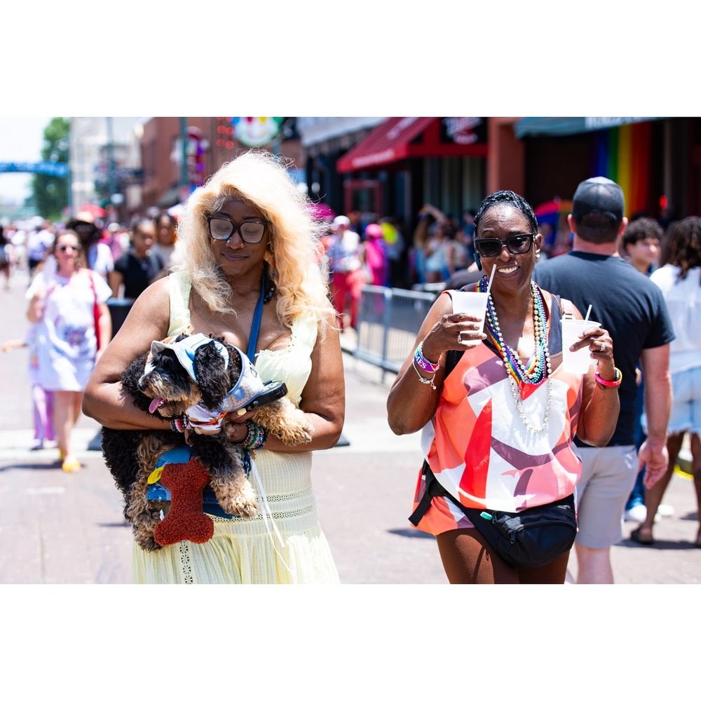 Color. Sunny Day. Looking leftward, down the middle of a brick-lined pedestrian avenue. Shops line the street in the background, receding from right to left. A slight crowd, in the background, ambling about. In the foreground, facing the viewer: two people, side-by-side. The person on the right has long, full, blond hair and brown skin, glasses, and a yellow dress. Their face is turned down to the small dog that they hold in their arms. The person on the right has short, black hair and brown skin and wears sunglasses, a colorful shirt, and several strings of beads around their neck. They smile at the viewer behind two plastic cups that their hands hold up.