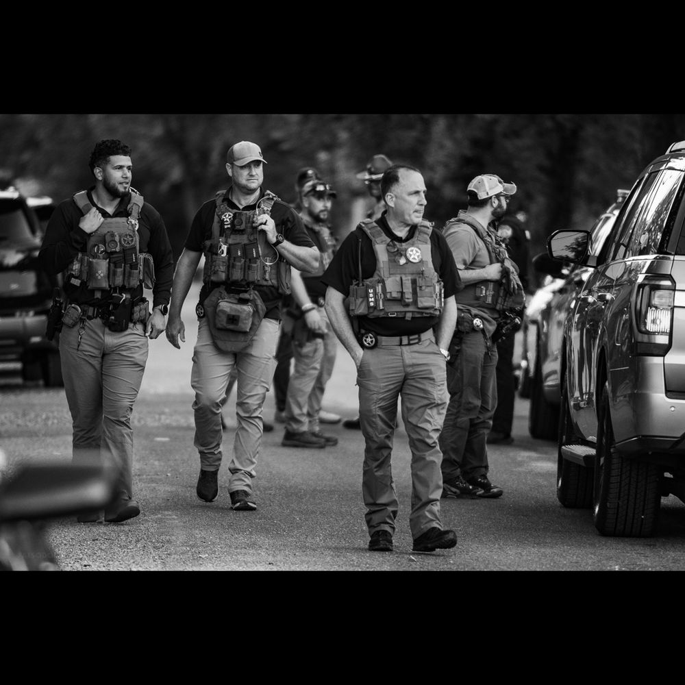 B&W. Day. Looking down a tree-lined suburban street. Three armed U.S. marshals in tactical vests walk down the middle of the street, toward the viewer. Several more men in such vests stand with a TN state trooper, in the background.