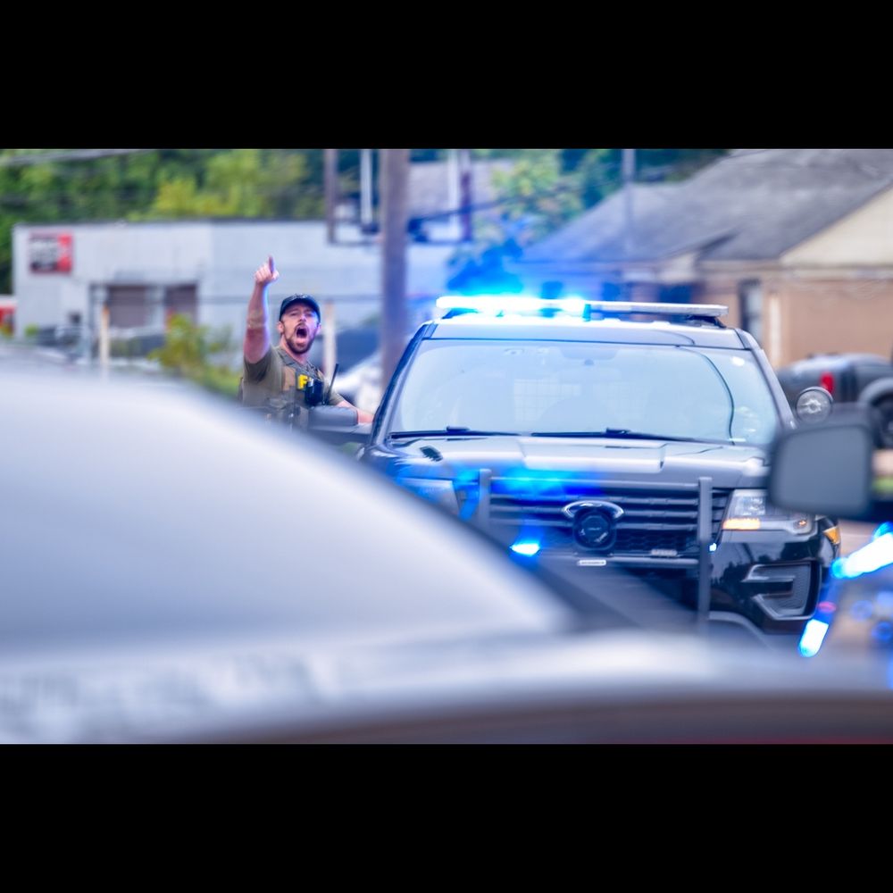 Color. Day. At a distance, the front end of a police SUV with blue lights illuminated on top. At the SUV's left, a man in a cap and a bulky vest on which a yellow "F" (of FBI) is visible, shouts angrily toward the viewer, his right arm in the air and his index finger extended.