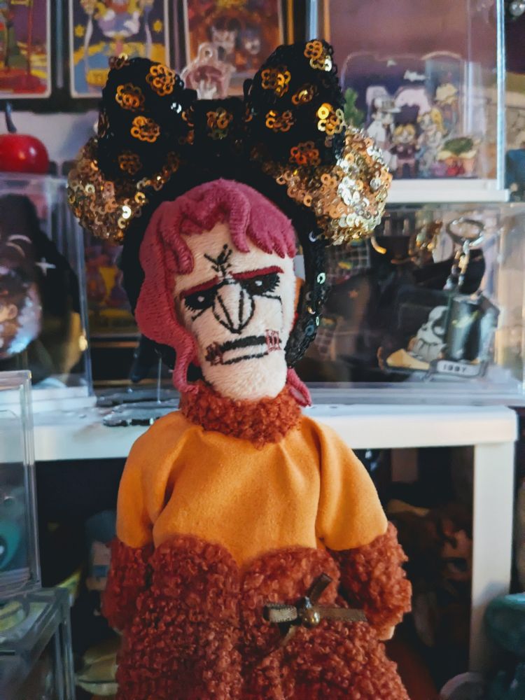 A soulless Kakyoin Noriaki doll from the JoJo's Bizarre Adventure franchise wearing an orange dress and wearing a gold Minnie Mouse ears headband with a black and gold bow