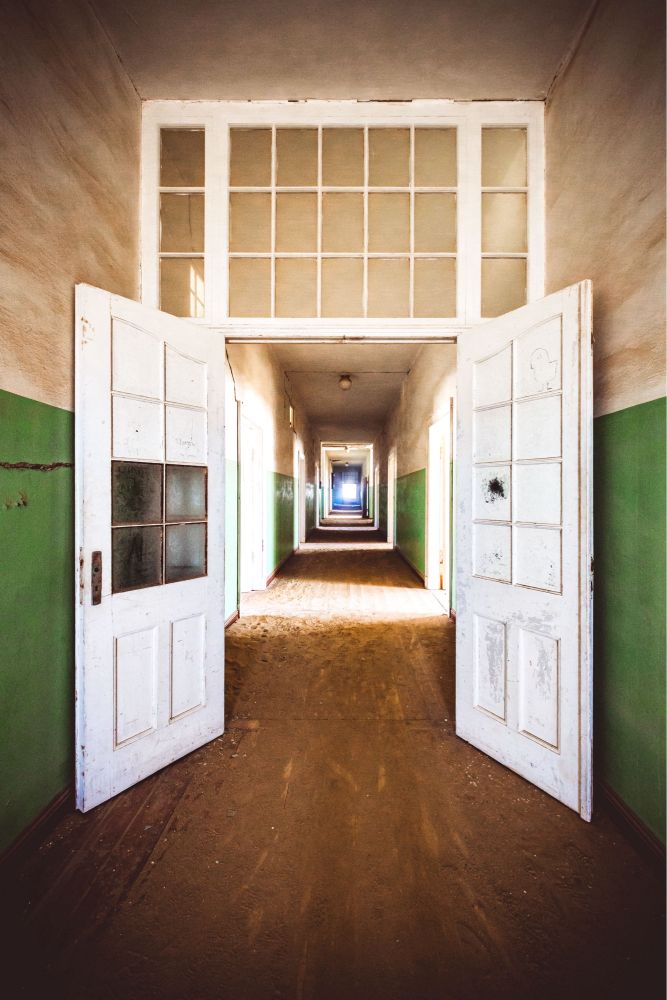 Wir blicken durch eine weiße Doppeltür einen sehr langen Flur hinunter. Ein Ende ist nicht zu sehen, nur helles Licht. Hinter der Doppeltür ist zu erkennen, dass vom Flur links und rechts Räume angehen, da durch Türöffnungen Licht in den Flur scheint. Die Wände links und rechts sind zur Hälfte grün (unten) und zur Hälfte cremefarben gestrichen. Der Holzboden ist mit Sand und Dreck bedeckt.