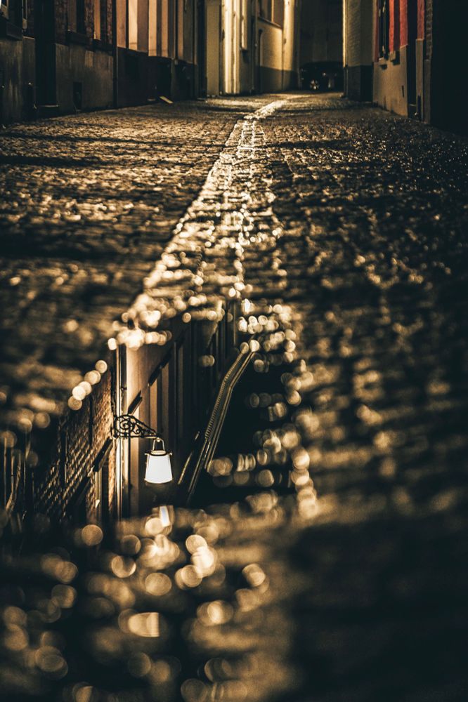 Enge, nächtliche Gasse mit nassem Kopfsteinpflaster, das das warme Licht einer Straßenlaterne reflektiert. Die Stimmung ist ruhig und atmosphärisch, mit golden schimmernden Reflexen auf dem Boden.