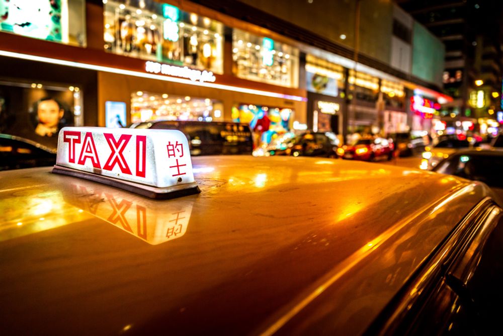 Nahaufnahme eines beleuchteten Taxi-Schilds mit der Aufschrift ‚TAXI‘ in Rot und chinesischen Schriftzeichen. Das Schild spiegelt sich auf dem Autodach, im Hintergrund leuchtet eine belebte Straße mit vielen Lichtern und Schaufenstern bei Nacht.