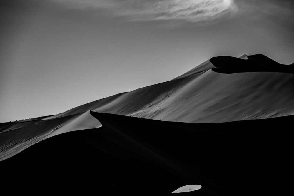Schwarz-Weiß-Fotografie einer weitläufigen Dünenlandschaft. Sanft geschwungene Linien aus Licht und Schatten betonen die Form der Sanddünen unter klarem Himmel. Die Szene wirkt ruhig und minimalistisch.