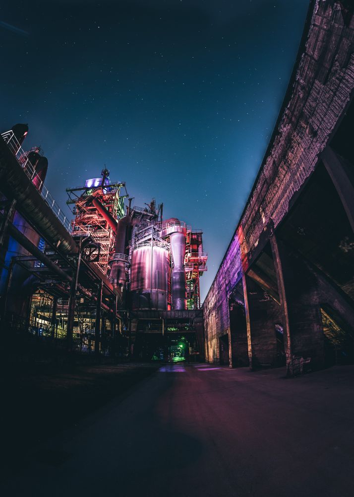 Nächtliche Ansicht eines Industriekomplexes mit großen metallischen Strukturen, Rohren und Türmen, die in kräftigen Farben wie Violett, Rot, Grün und Blau beleuchtet sind. Der dunkle Himmel ist mit Sternen übersät. Die Perspektive zeigt die Szene vom Boden aus, mit einer gebogenen Wand oder Überführung rechts im Bild, die Tiefe und Rahmen verleiht.