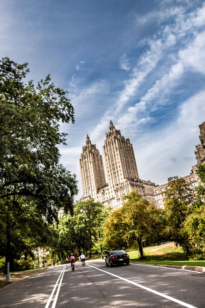 Bildbeschreibung:

Das Foto zeigt eine Szene im Central Park in New York City, aufgenommen bei Tageslicht mit Blick auf eine breite Straße, die durch den Park führt. Im Hintergrund ragen zwei markante Hochhäuser in den Himmel.

Vordergrund und Straße:

• Eine asphaltierte Straße verläuft leicht diagonal von unten links nach oben rechts.
• Die Straße ist mit zwei weißen Linien markiert, die eine Fahrspur für Radfahrer oder Jogger abgrenzen.
• Auf der Straße sind mehrere Menschen unterwegs: Zwei Radfahrer fahren nebeneinander, dahinter gehen zwei Personen zu Fuß, eine davon mit einem pinken Oberteil.
• Ein schwarzes Auto fährt ebenfalls auf der Straße, weiter hinten.


Umgebung und Natur:

• Links und rechts der Straße stehen viele Bäume mit grünen und leicht herbstlich gefärbten Blättern.
• Die Bäume sind hoch und dicht, einige werfen Schatten auf die Straße.
• Der Park wirkt gepflegt und ruhig, mit Rasenflächen und vereinzelten Büschen.


Hintergrund und Gebäude:

• Im Hintergrund ragen zwei identische, sehr hohe Gebäude mit spitzen Türmen in den Himmel. Sie sind im Stil des Art Déco gebaut und haben eine helle Sandsteinfarbe.
• Die Gebäude sind Teil der berühmten “San Remo Apartments”, die direkt am Rand des Central Parks stehen.
• Weitere kleinere Gebäude sind rechts im Bild zu sehen, teils von Bäumen verdeckt.


Himmel und Lichtstimmung:

• Der Himmel ist blau mit langen, weißen Schleierwolken, die sich dynamisch über das Bild ziehen.
• Die Lichtstimmung ist hell und freundlich, die Sonne scheint von rechts und beleuchtet die Gebäude und Bäume warm.

