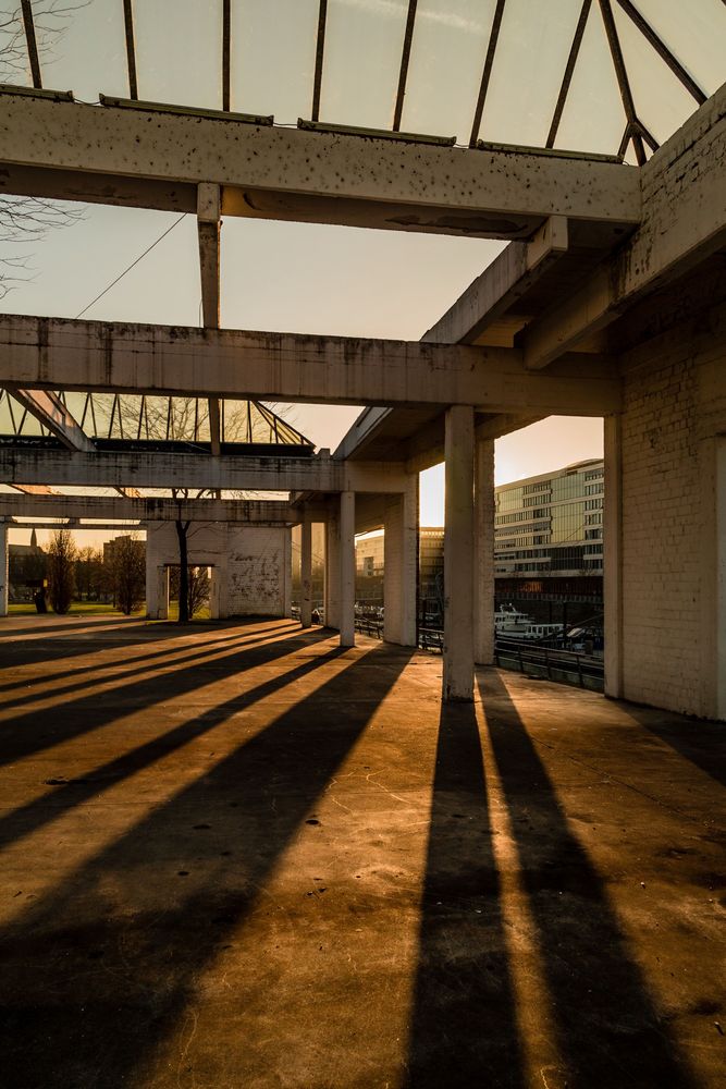 Offene Betonhalle bei Sonnenuntergang mit langen Schatten der Säulen auf dem Boden. Teilweise verfallene Dachstruktur mit Glasflächen. Im Hintergrund moderne Gebäude und ein Wasserkanal mit Booten. Ruhige, warme Lichtstimmung.