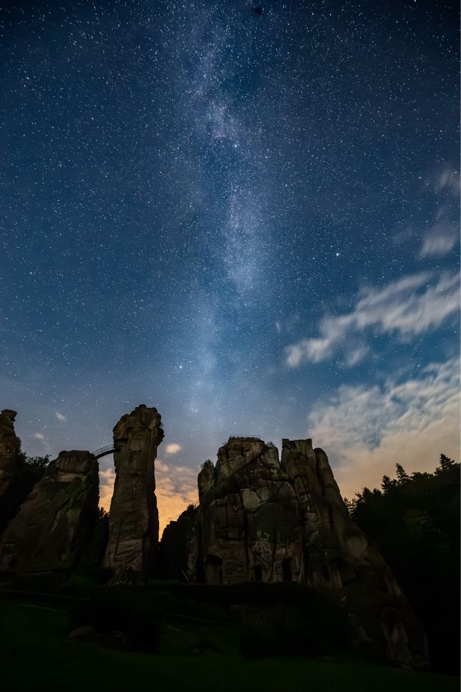 Nachtaufnahme. Im Vordergrund liegt eine Sandstein Formation im Schatten/dunkeln. Der unterste Teil des Bildes ist fast gänzlich schwarz.
Darüber strahlt der nächtliche Sternenhimmel in tiefem Blau. Ein paar wenige Wolkenfetzen sind zu sehen. In der Mitte ist die Milchstraße auszumachen.
