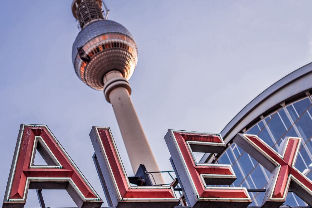 Rotes Werbeschild von schräg unterhalb fotografiert. Zu lesen ist: ALEX
Im Hintergrund ist ein Fernsehturm im späten Licht zu sehen.