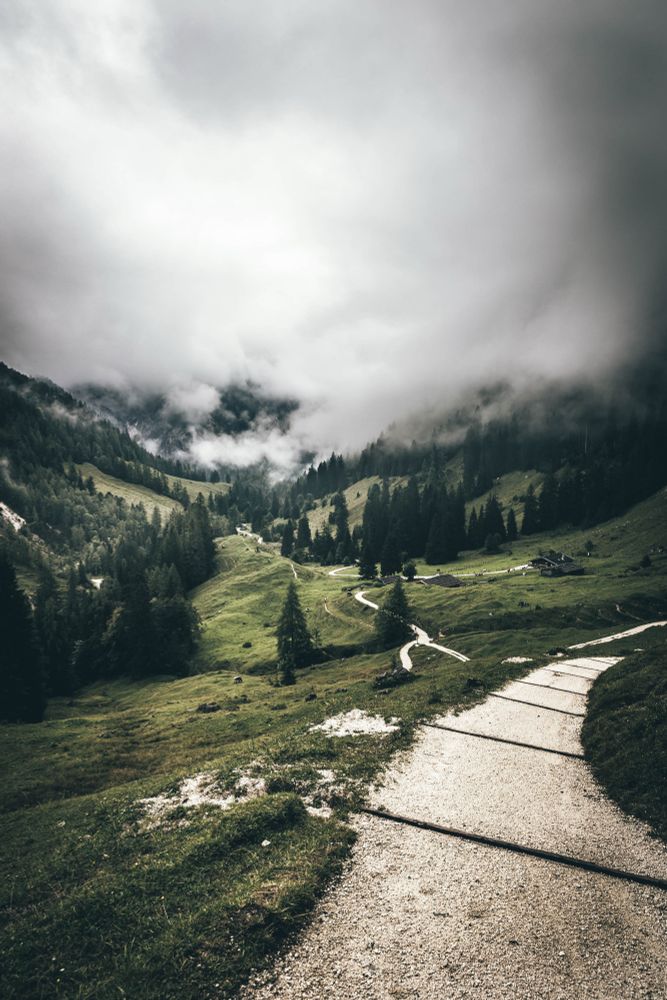 Berglandschaft mit einem gewundenen Kiesweg, der durch grüne Wiesen und zwischen dunklen Tannen verläuft. Dichte Wolken hängen tief in den Tälern und verdecken teilweise die Berghänge, wodurch eine mystische, neblige Stimmung entsteht.