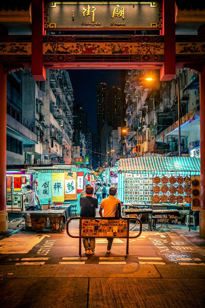 Blick durch ein verziertes Tor mit der Aufschrift ‚Temple Street‘ auf einen belebten Nachtmarkt in einer engen Stadtstraße. Unter farbigen Lichtern und Marktständen mit Bannern, Lampen und Waren stehen zwei Personen mit dem Rücken zur Kamera und schauen in die lebendige Szenerie.