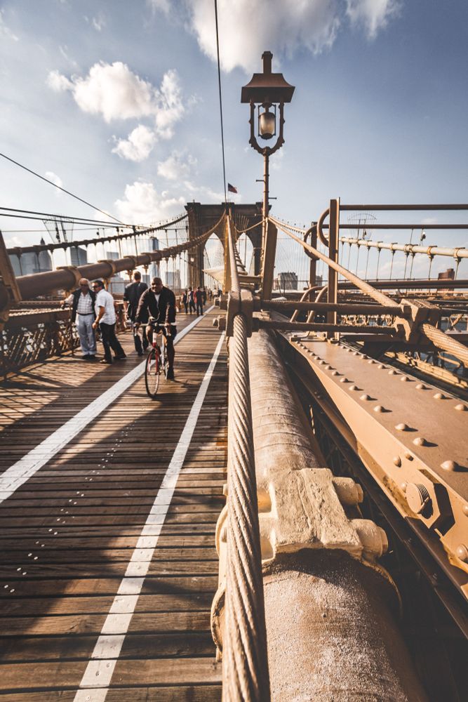 Bildbeschreibung:

Das Foto zeigt die Brooklyn Bridge in New York City aus der Perspektive eines Fußgängers, der sich auf dem Holzsteg der Brücke befindet und in Richtung der steinernen Brückenpfeiler blickt. Die Aufnahme ist bei Tageslicht entstanden, vermutlich am Nachmittag, da die Sonne warmes Licht auf die Szene wirft und lange Schatten erzeugt.

Vordergrund:

• Der Holzsteg der Brücke ist in zwei Spuren unterteilt: links für Fußgänger, rechts für Radfahrer. Die Spuren sind durch weiße Linien markiert.
• Ein dicker Stahlseilzug verläuft diagonal von unten rechts nach oben links und dominiert den Vordergrund. Er ist mit rostbraunen Metallverbindungen befestigt.
• Ein Laternenpfahl mit einem nostalgischen Design steht mittig im Bild und ragt in den Himmel.

Menschen:

• Mehrere Personen sind auf dem Steg unterwegs. Links gehen Fußgänger, darunter ein Mann mit weißem Hemd und eine Frau mit Sonnenbrille.
• In der Mitte fährt ein Radfahrer mit dunkler Kleidung direkt auf die Kamera zu.
• Die Menschen wirken entspannt, einige unterhalten sich, andere fotografieren.


Hintergrund:

• Die markanten steinernen Türme der Brooklyn Bridge mit ihren gotischen Bögen sind deutlich zu sehen.
• Über die Brücke verlaufen zahlreiche Stahlseile, die sich fächerartig von den Türmen ausbreiten.
• Im Hintergrund sind die Umrisse von Hochhäusern Manhattans zu erkennen, leicht verschwommen durch die Perspektive und das Licht.


Himmel und Atmosphäre:

• Der Himmel ist blau mit einigen weißen, bauschigen Wolken.
• Die Lichtstimmung ist warm und golden, was dem Bild eine freundliche und lebendige Atmosphäre verleiht.