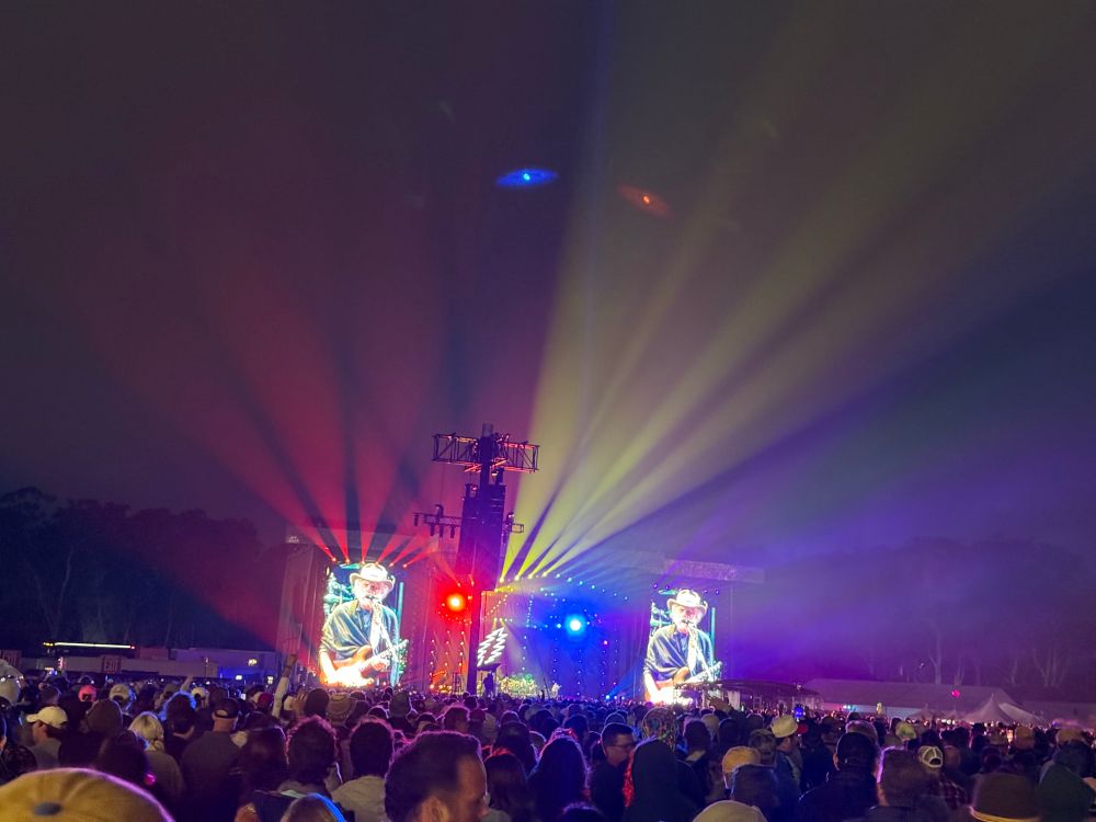 A stage seen from a distance. There is a light fog and the stage lights are projecting psychedelic rays into the night sky