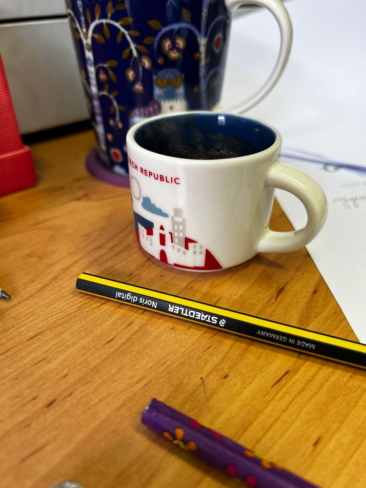 Hrníček na espresso (s nápisem Czech republic) na stole, za ním větší modrý hrnek s obrázky, kolem binec - tužky, papíry. 