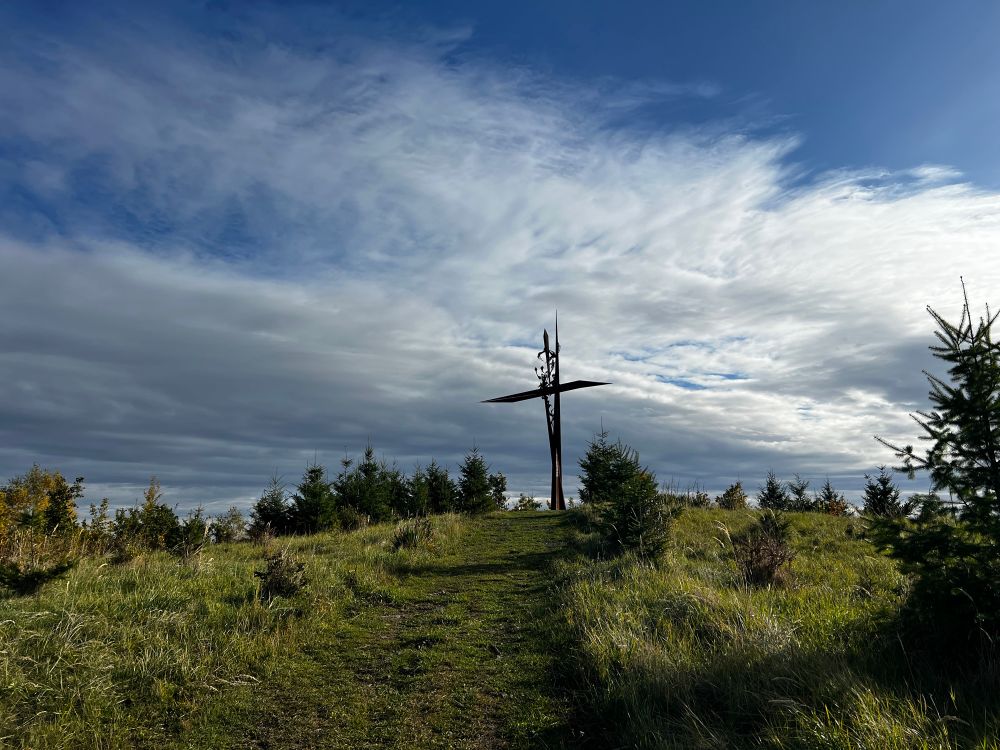 Kovový umělecký kříž na kopci s bílými oblaky v pozadí 