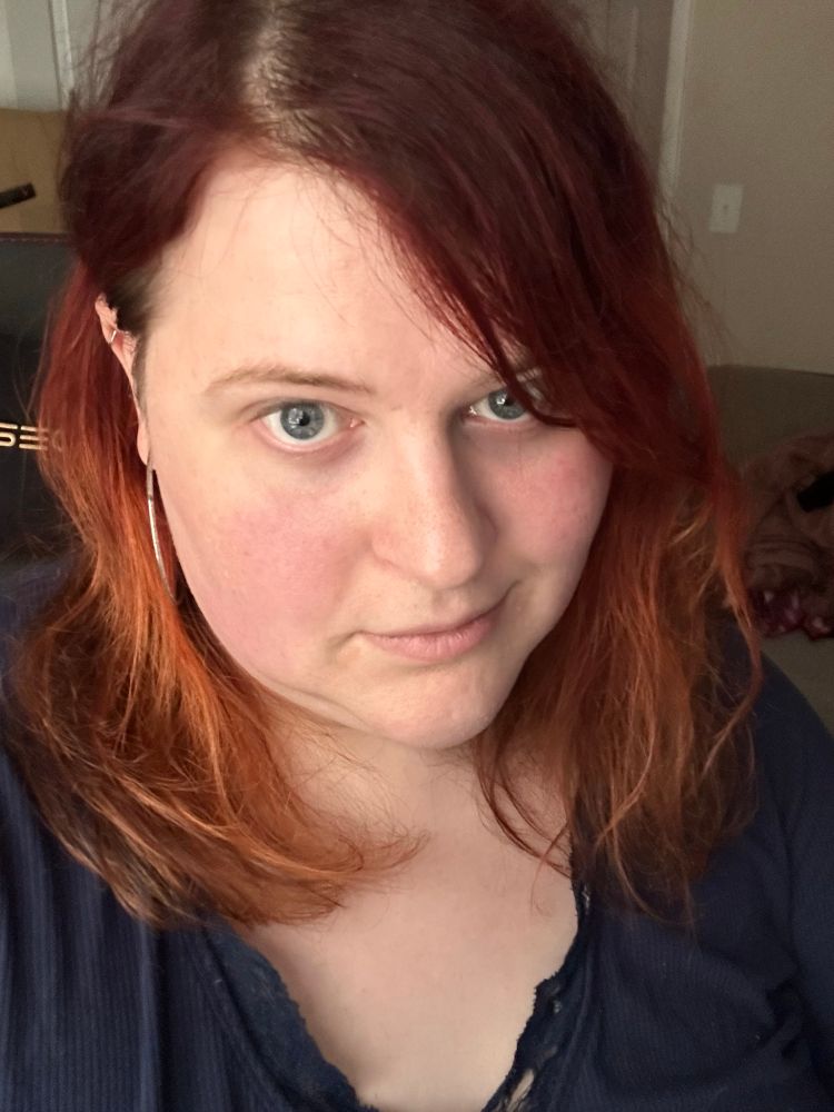 Kat, a woman with red hair and a sardinic expression working from home wearing no makeup and no bra