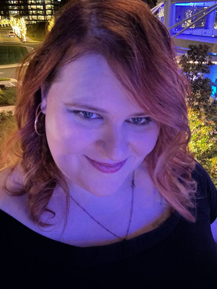 Kay, a redheaded woman with curled hair wearing a black off the shoulder dress, photographed on a balcony at night