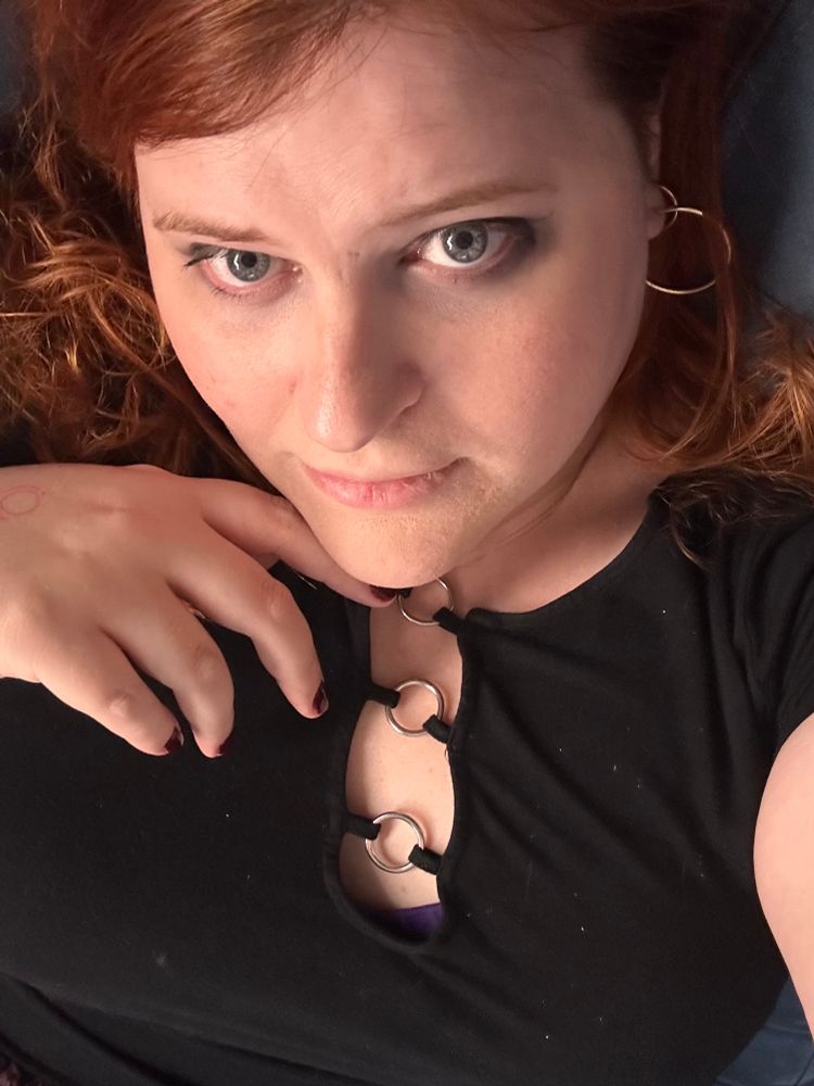 Kat, a red haired woman with intense blue eyes, laying down on the couch and looking up. The photo is taken from the angle of someone above her looking down