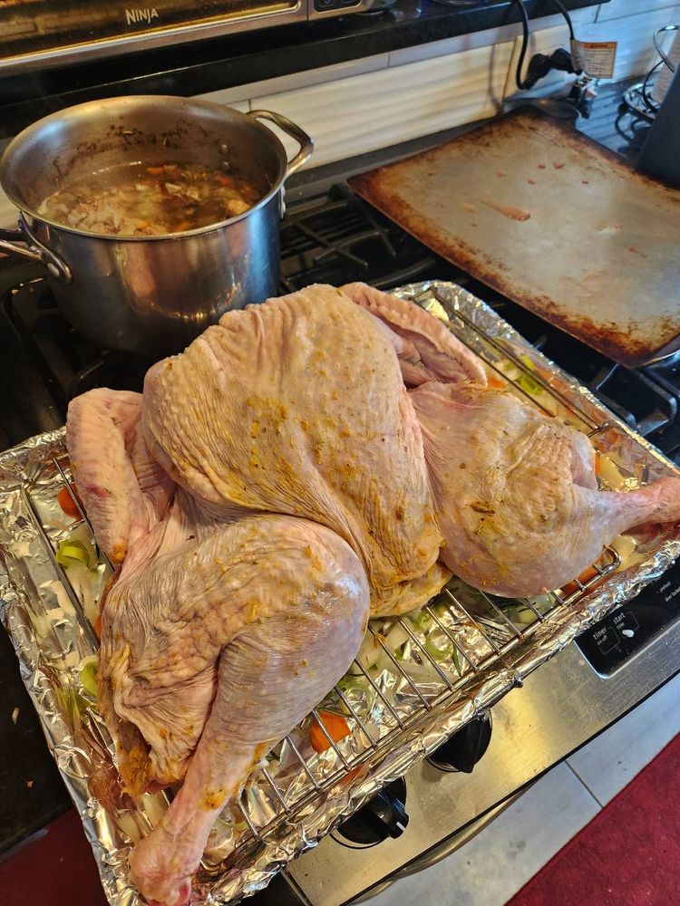 A butterflied turkey sitting on a cooking rack over vegetables, with a stock pot of ingredients on the back burner.
