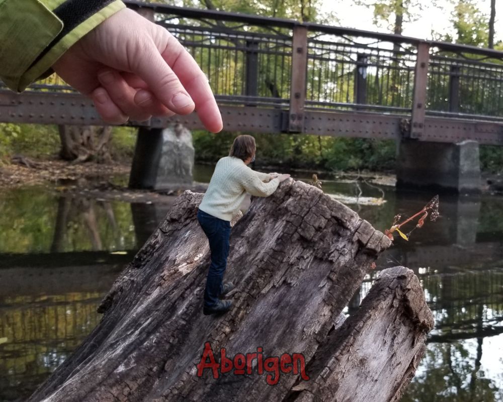 A tiny man in a fisherman's sweater climbs an angled stump overlooking a creek. A gigantic woman's hand reaches down to pluck him up.