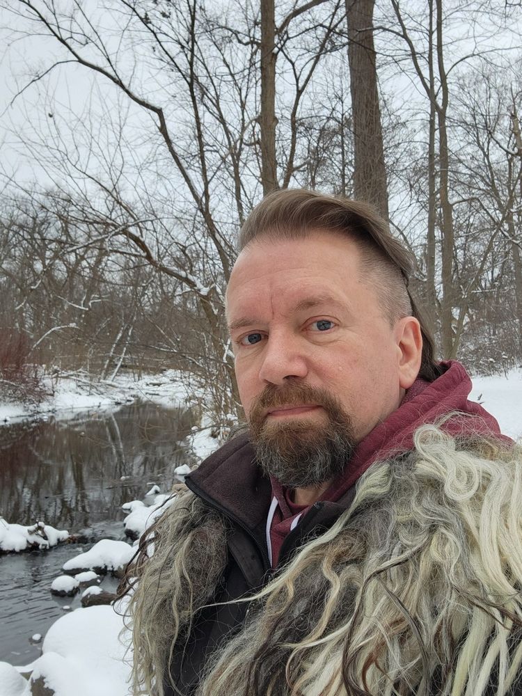 Aborigen stands in winter gear before a placid creek lined by snowy banks and barren trees.