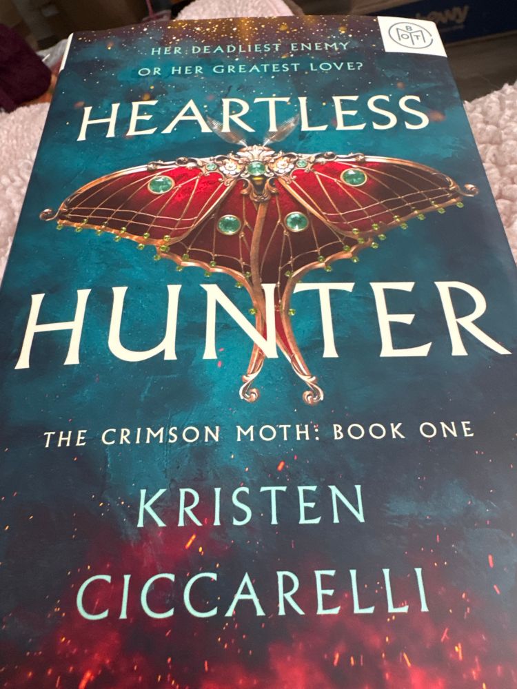 The book cover for the novel “ Heartless Hunter” by Kristen Ciccarelli. The cover had a beautiful read, gold and emerald green butterfly with flames below it and stars above it. 