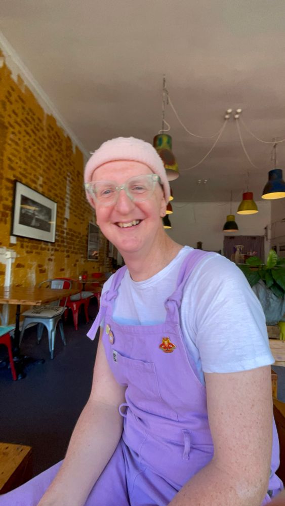 A pale white woman (me) in lilac overalls with a white t-shirt underneath and a pastel pink beanie with green plastic glasses. She's smiling though clearly reluctant to be in front of the camera