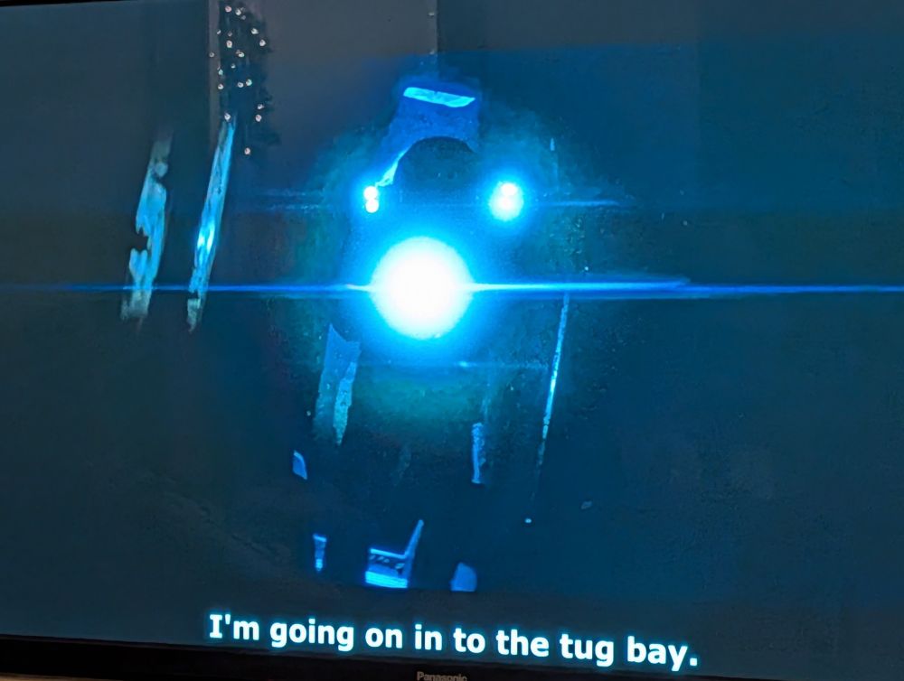 Man in spacesuit pointing a spotlight directly at the camera, subtitle reads "I'm going on in to the tug bay."