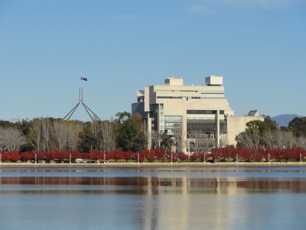 The High Court of Australia, on the autumn shores of Lake Burley Griffin, with the spire of the Parliament of Australia in the background