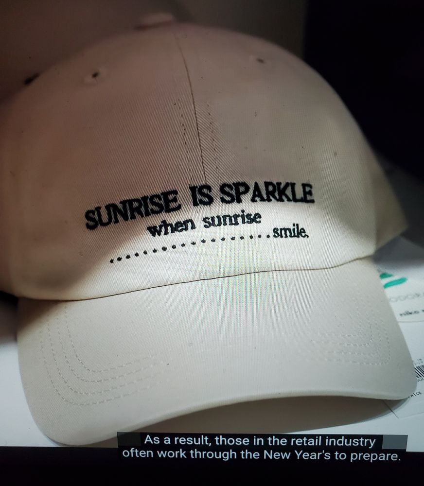 A white dad hat with black, serif text on the front that reads, "SUNRISE IS SPARKLE when sunrise......................smile." The subtitle overlaid on the bottom of the image also reads, "As a result, those in the retail industry often work through the New Year's to prepare."