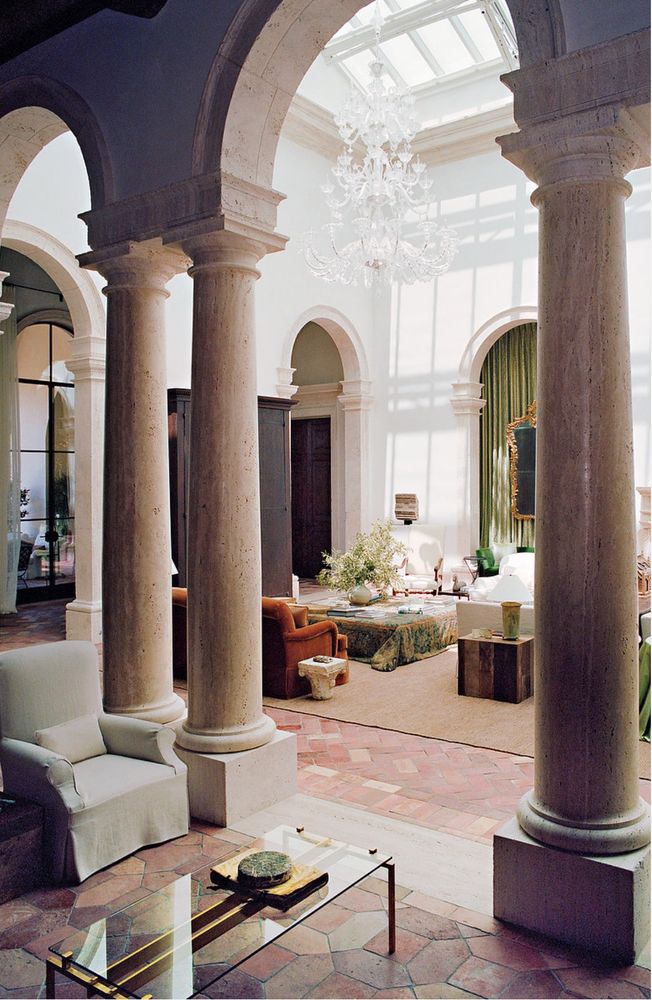 Double-height living room, with clear Venetian influence, seen through a stone colonnade with brick floors, upholstered furniture and light streaming from above, along with a Venetian glass chandelier 