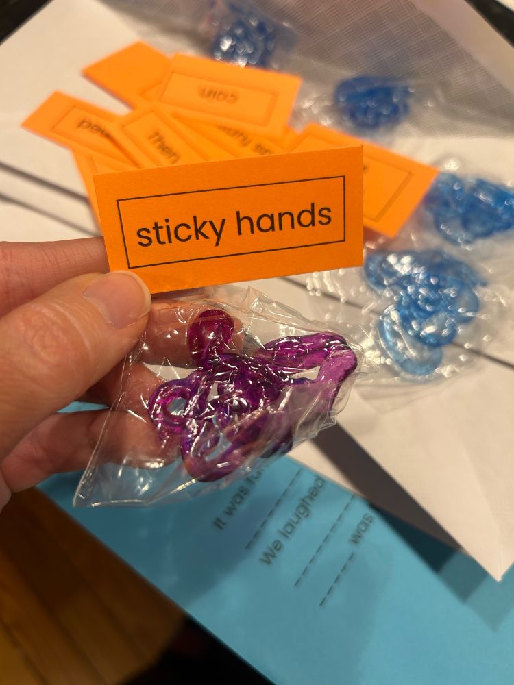 A small clear bag that has a purple sticky hand toy inside. Above the bag is an orange piece of paper that reads, “sticky hands.” There are other small pieces of orange paper, white envelopes, and blue pieces of paper in the background.