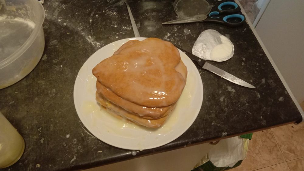 a cake, stacked out of centimeter-thick brown baked-dough layers, covered and leaking with cream sitting atop a round white plate. the top layer of the cake is shaped like a heart. traces of what looks like mysterious white powder (flour) and scattered utensils litter the battlefield that is this kitchen counter, glorifying the cake's creation