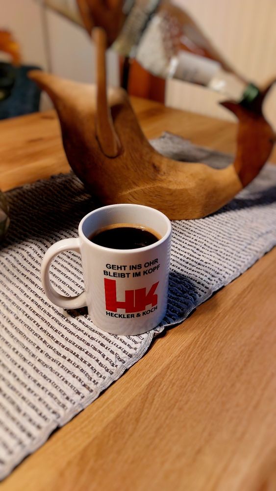 Eine Tasse Kaffee mit der Aufschrift:Geht ins Ohr,bleibt im Kopf.Darunter das Logo der Firma Heckler & Koch