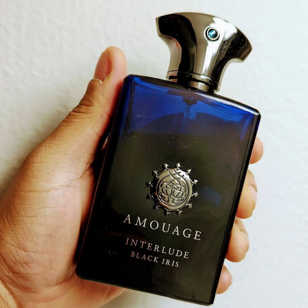 White background, hand holding a dark-blue bottle with a silver cap. Lettering: Amouage Interlude: Black Iris