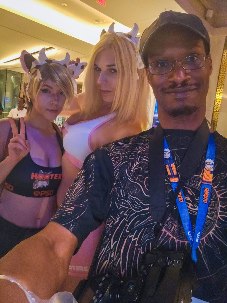 Bsky user C0splaygirlz dressed as cows, along with me at the convention Otakon 2025. C0splaygirlz are wearing cow ears and Hooters outfits,  and one of them is posing making a peace sign with her fingers 