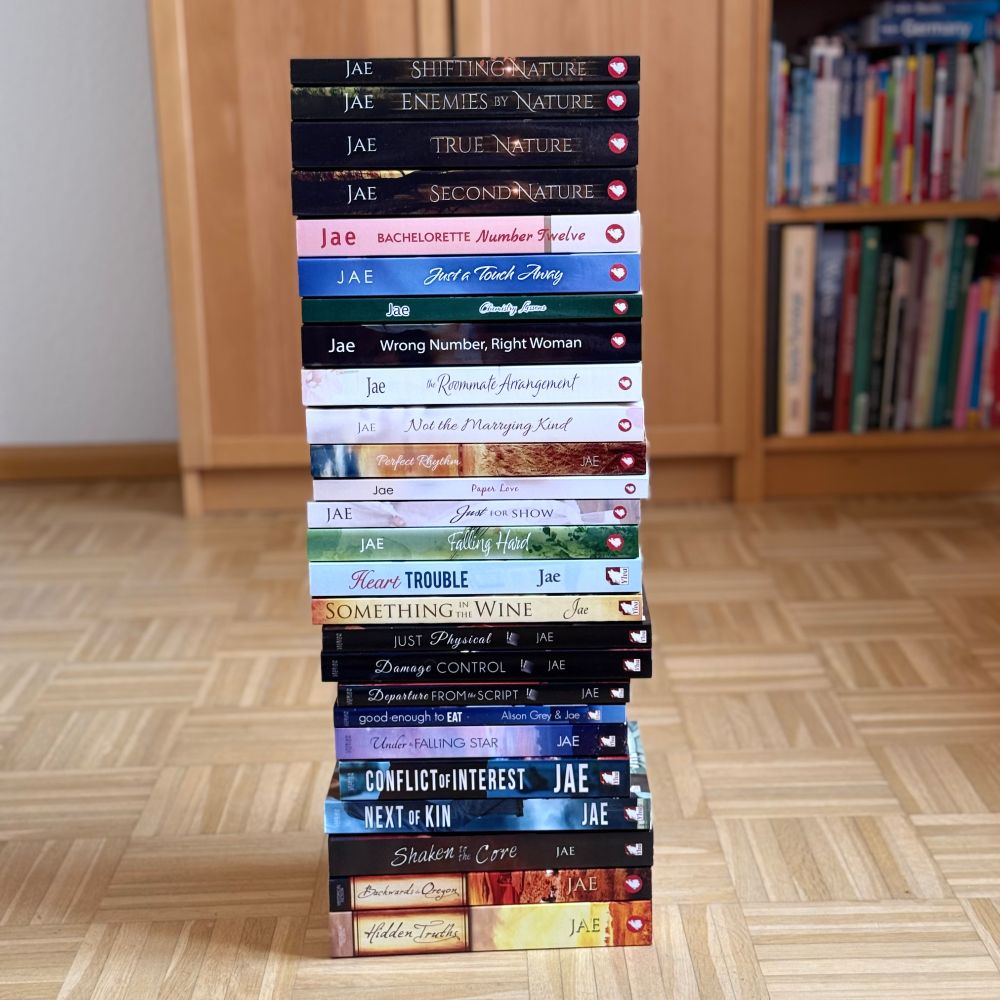 Depicted is a stack of Jae's 26 novels, on a hardwood floor in front of a bookshelf. 