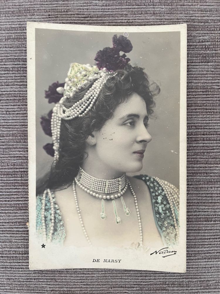 Jeanne de Mary by Nadar. Bust profile photo of white woman draped in many strands of pearls including a choker, around her neck, over her shoulder and in her hair which is half up. Low cut bodice tinted aqua seems covered in sequins. Pink lipstick and burgundy flowers in her hair; chest faintly skin tone. Postcard otherwise B&W