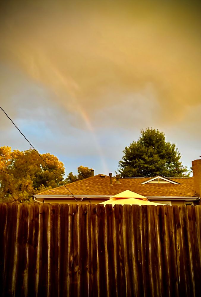 Shot of a cloudy sky over a house and trees, and a wooden fence in the foreground. Ripping from the clouds into the roof of the house, is a faint rainbow, almost aligned with dipping clouds to look like a forming tornado.
