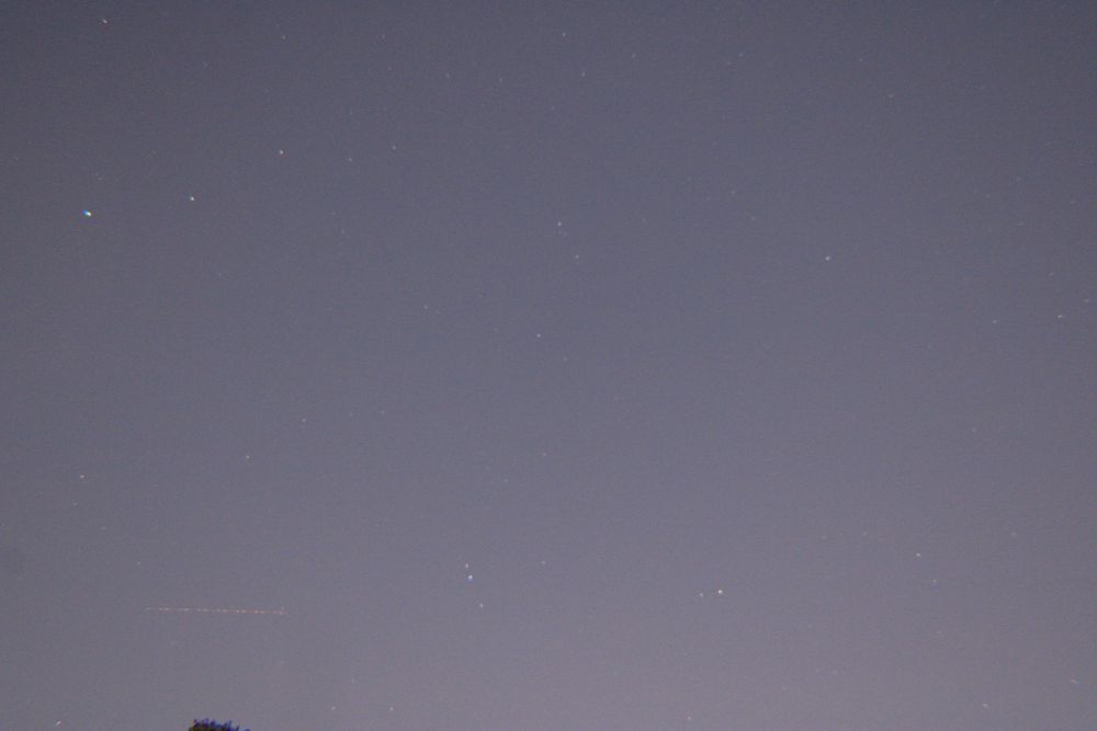 An expanse of night sky, light pollution casts the sky a dull grey, but zooming in reveals several bright stars.
In the lower left, a line is being formed by some sort of object in flight, a plane, satellite, meteor—unknown.