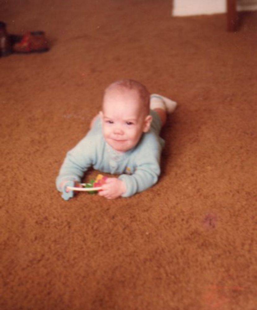 A baby boy—infant author to be, J.D. Buffington—wearing a blue onesie and white socks. He is holding a classic, plastic key teething toy. He is looking straight into camera with a smirk as if he knows something funny. He lays, head and arms toward us, back and legs behind him (obscured by his big, baby head), on a vast expanse of brown carpet—so 70’s—he is smirking as if he either knows something funny or has just shit his diaper. This IS a photo of a baby.