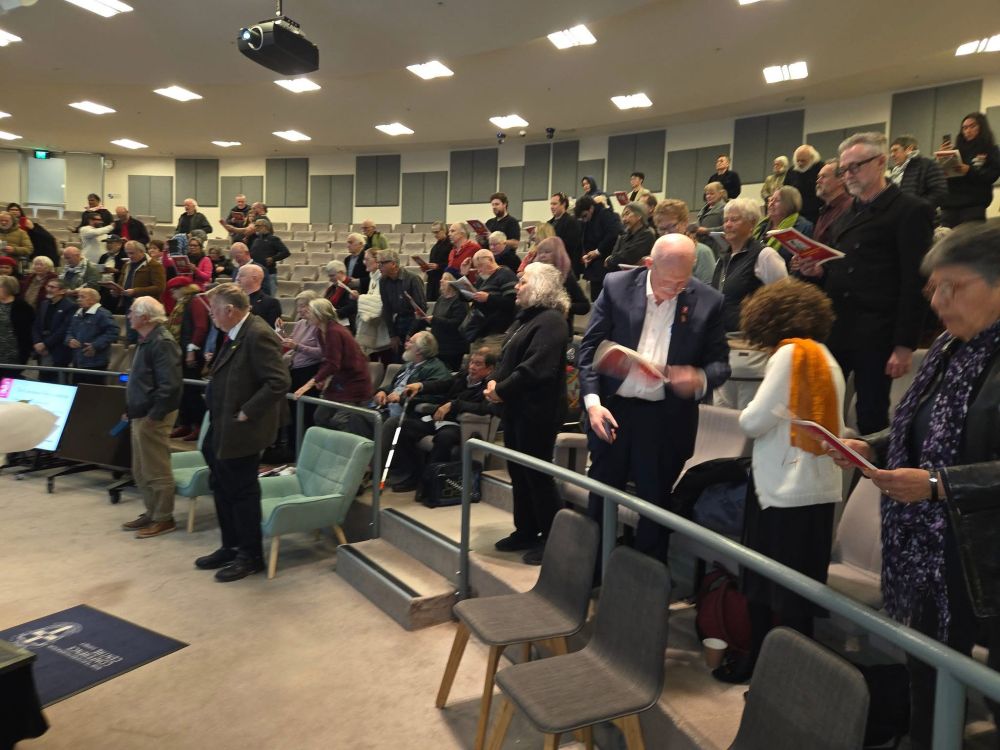 An auditorium full of people standing up at Hannah Middleton's memorial: Denis Doherty at the front