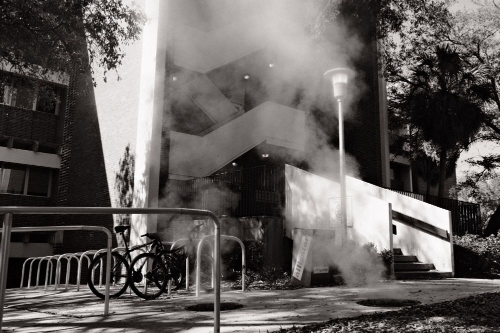 A black and white picture of a staircase behind two manholes that are emitting steam. 